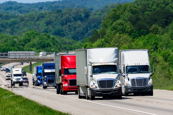 Multiple semi-trucks driving side-by-side on a highway surrounded by trees and rolling hills under a clear sky.