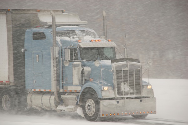 A blue semi-truck driving through a heavy, blowing snowstorm on a white, snow-covered Indiana highway.